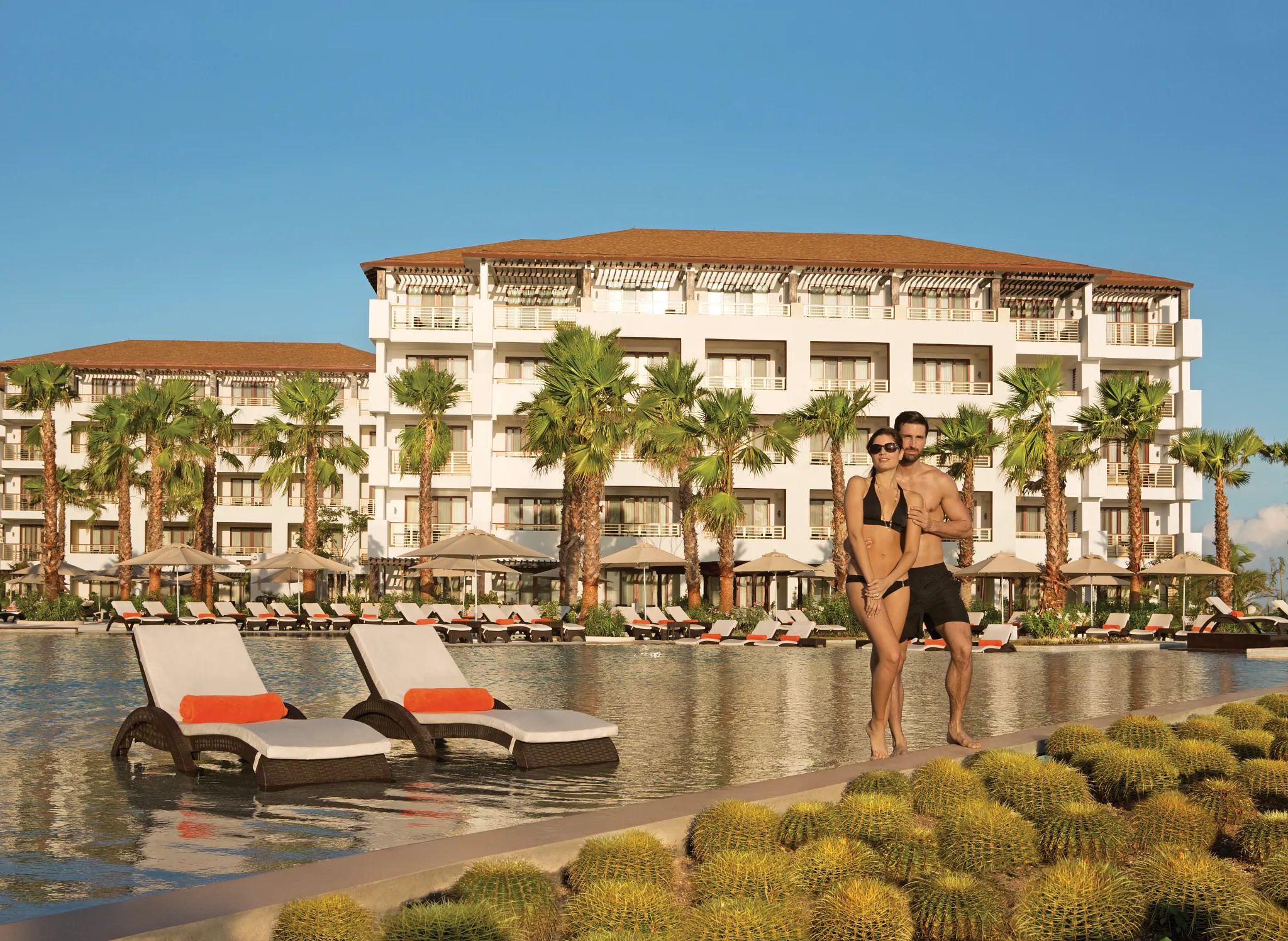 Couple standing beside the resort pool with palm trees and the resort buildings behind them at Secrets Playa Mujeres Golf & Spa Resort.